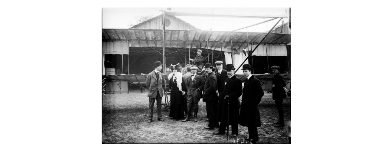 Mario Nunes Vais, Gruppo di spettatori fotografati a Campo di Marte in occasione della I Dimostrazione di volo - alle loro spalle si osserva un biplano con a bordo un aviatore e, sul fondo, gli hangars, 1910, gelatina ai sali d'argento, F034476